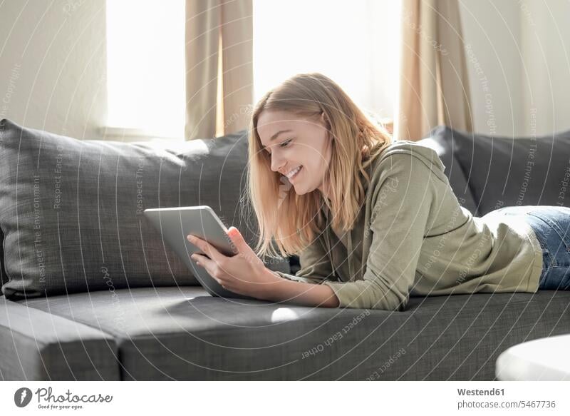Porträt einer lächelnden jungen Frau, die zu Hause auf der Couch liegt und ein digitales Tablet benutzt Leute Menschen People Person Personen Alleinstehende