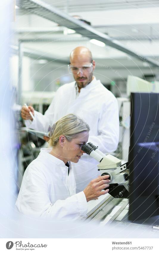 Eine Wissenschaftlerin forscht, während ein männlicher Kollege im Labor ein Mikroskop betrachtet Farbaufnahme Farbe Farbfoto Farbphoto Innenaufnahme