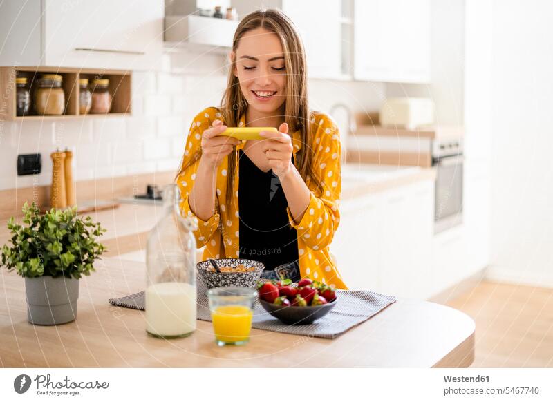 Glückliche junge Frau benutzt Mobiltelefon und frühstückt zu Hause in der Küche Leute Menschen People Person Personen Alleinstehende Alleinstehender Singles