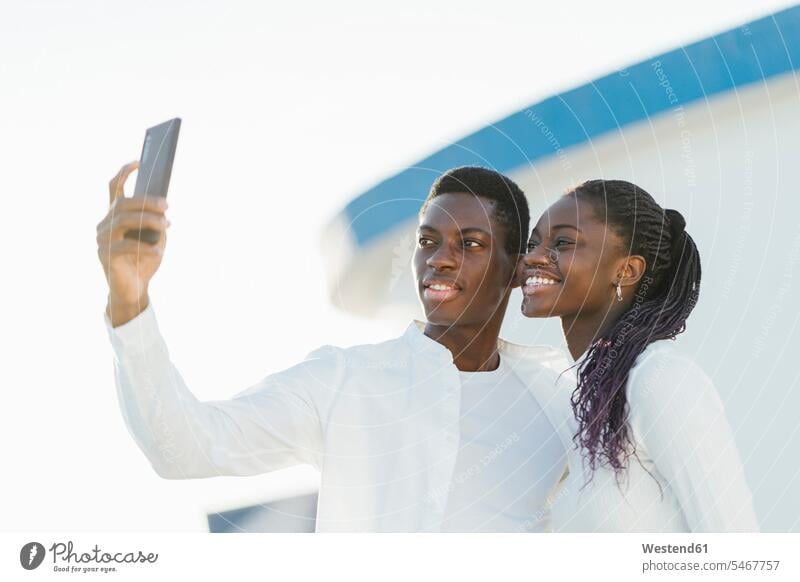Lächelndes Paar nimmt Selfie auf mit dem Handy vor weißer Wand Farbaufnahme Farbe Farbfoto Farbphoto Außenaufnahme außen draußen im Freien Tag