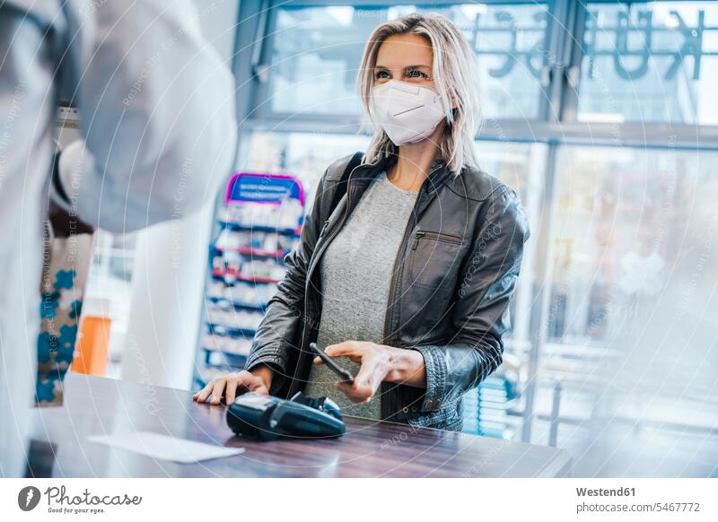 Weibliche Kundin mit Gesichtsmaske beim Bezahlen mit dem Smartphone an der Kasse eines Drogeriemarktes Farbaufnahme Farbe Farbfoto Farbphoto Innenaufnahme