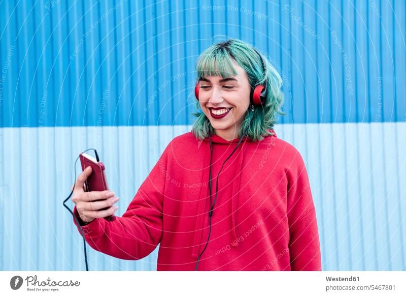 Porträt einer lachenden jungen Frau mit blau gefärbten Haaren und Kopfhörern, die ein Selfie mit ihrem Mobiltelefon macht blaue blauer blaues faerben färben