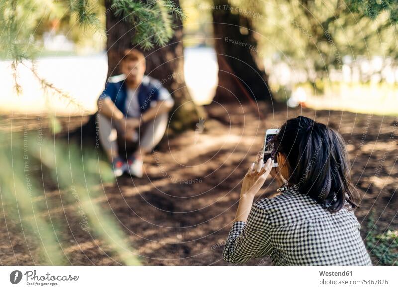 Junge Frau macht Handyfoto von ihrem Freund unter einem Baum Telekommunikation telefonieren Handies Handys Mobiltelefon Mobiltelefone entspannen relaxen sitzend