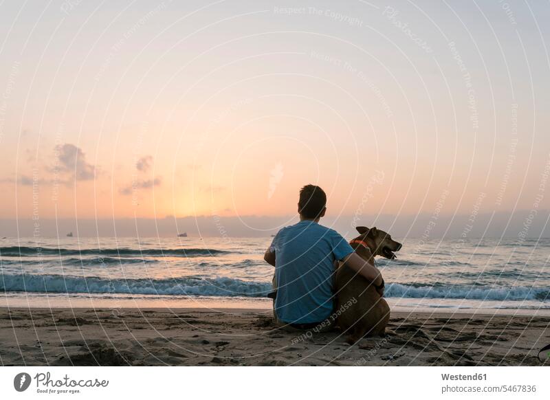 Mann sitzt mit seinem Hund und genießt den Sonnenaufgang am Strand Farbaufnahme Farbe Farbfoto Farbphoto Außenaufnahme außen draußen im Freien Freizeitkleidung