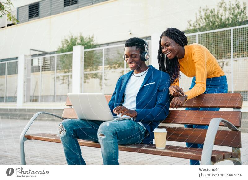 Glückliches Paar schaut auf Laptop auf Bank Farbaufnahme Farbe Farbfoto Farbphoto Außenaufnahme außen draußen im Freien Tag Tageslichtaufnahme