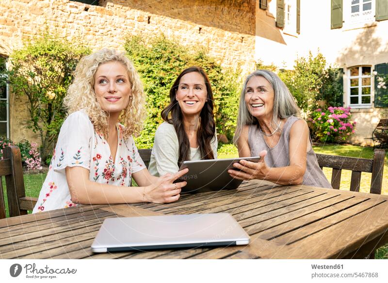 Drei Frauen unterschiedlichen Alters sitzen am Gartentisch und benutzen ein Tablet glücklich Glück glücklich sein glücklichsein Terrassentisch Tablet Computer
