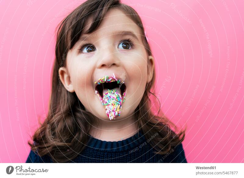 Porträt eines glücklichen kleinen Mädchens mit Zuckergranulat auf Lippen und Zunge vor rosa Hintergrund begeistert Enthusiasmus enthusiastisch Überschwang