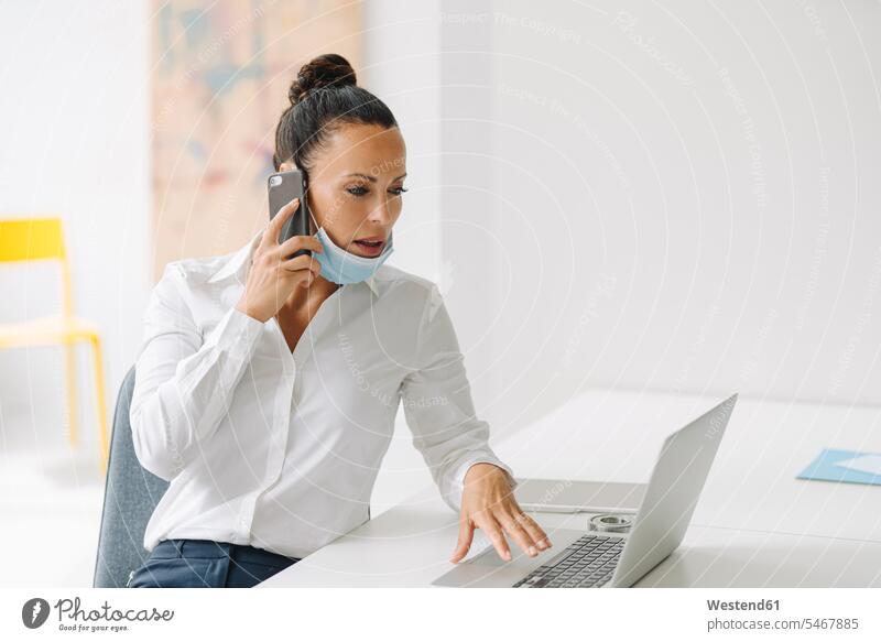 Geschäftsfrau mit Maske, die über ein Smartphone telefoniert, während sie den Laptop auf dem Schreibtisch im Heimbüro benutzt Farbaufnahme Farbe Farbfoto