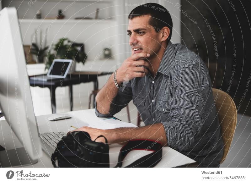 Junger Mann mit Kamera, der am Schreibtisch sitzt und einen Computer benutzt Fotoapparat Fotokamera sitzen sitzend Rechner Arbeitstisch Schreibtische Männer