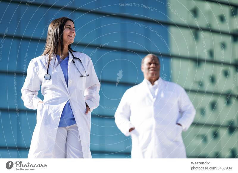 Zuversichtliche Ärzte stehen mit den Händen in den Taschen gegen das Krankenhaus Farbaufnahme Farbe Farbfoto Farbphoto Außenaufnahme außen draußen im Freien Tag