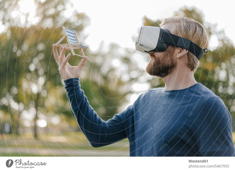 Mann im herbstlichen Park, der eine Virtual-Reality-Brille trägt und eine Miniatur-Strandliege betrachtet Strandliegen Männer männlich Virtual Reality Brille