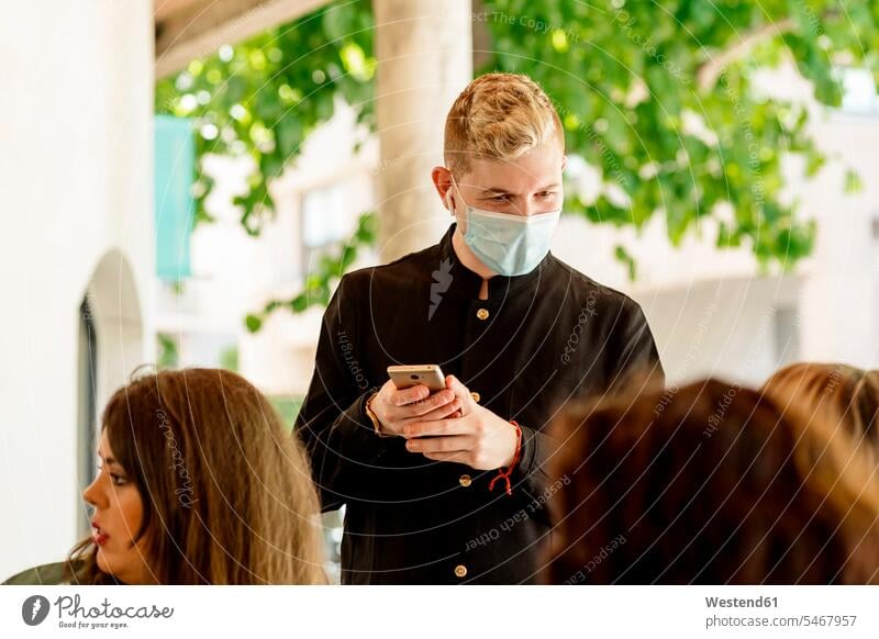 Junger Kellner hält Smartphone in der Hand, während er weibliche Kunden im Restaurant anschaut Farbaufnahme Farbe Farbfoto Farbphoto Spanien Innenaufnahme