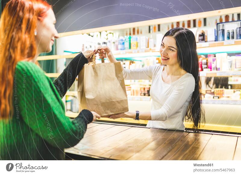 Lächelnde Verkäuferin, die in einem Kosmetikgeschäft eine Papiertüte an einen Kunden übergibt Papiertüten Geschäft Shop Laden Läden Geschäfte Shops
