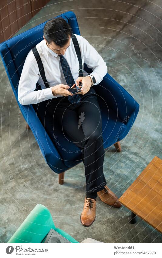 Draufsicht auf einen Geschäftsmann, der in einem blauen Sessel sitzt und ein Smartphone benutzt sitzen sitzend iPhone Smartphones Businessmann Businessmänner