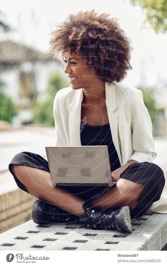 Modische junge Frau mit lockigem Haar sitzt auf Bank mit Laptop und lacht sitzen sitzend Notebook Laptops Notebooks Sitzbänke Bänke Sitzbank Locken