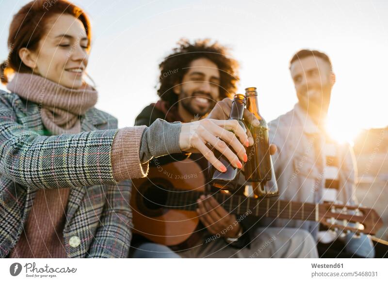 Drei glückliche Freunde mit Gitarre toasting Bierflaschen bei Sonnenuntergang sitzen sitzend sitzt Sonnenuntergänge anstoßen zuprosten anstossen Glück