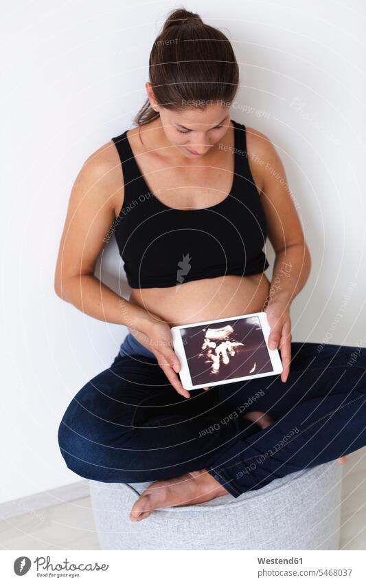 Junge schwangere Frau mit Ultraschallbild ihres ungeborenen Babys auf der Tablette Oberkoerper Oberkörper Torso Torsos Bäuche sitzend sitzt freuen Glück