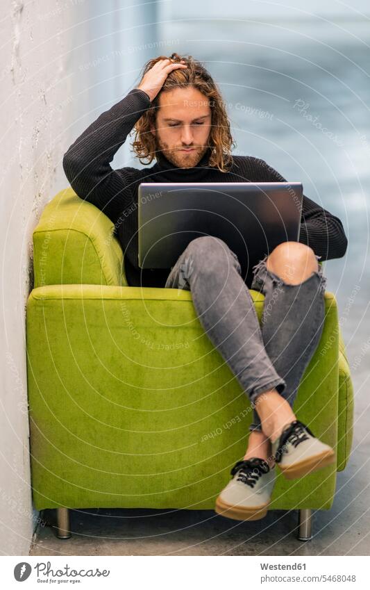 Junger Mann mit Hand in den Haaren, der einen Laptop benutzt, während er zu Hause auf einem Sessel sitzt Farbaufnahme Farbe Farbfoto Farbphoto Spanien