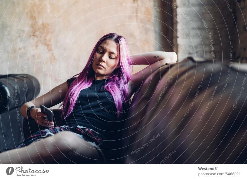 Junge Frau mit rosa Haaren sitzt mit einem Smartphone auf dem Sofa in einem Loft Couch Couches Liege Sofas sitzend entspannen relaxen entspanntheit relaxt