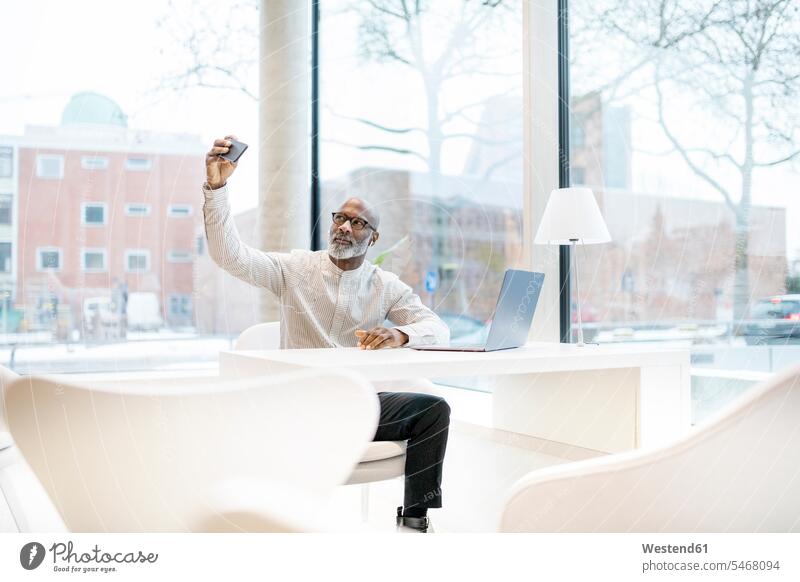 Porträt eines reifen Mannes mit Laptop, der am Schreibtisch sitzt und ein Selfie mit seinem Smartphone macht Geschäftsmann Businessmann Businessmänner
