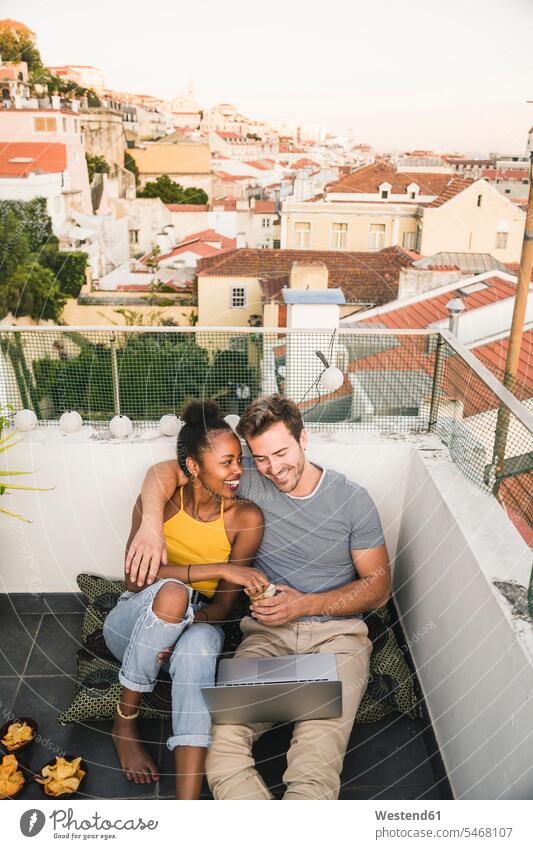 Glückliches junges Paar mit Laptop sitzt abends auf dem Dach, Lissabon, Portugal Polster Rechner Laptops Notebook Notebooks entspannen relaxen sitzend