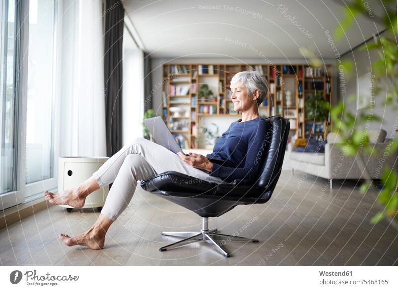 Aktive ältere Frau mit Laptop, während sie zu Hause auf einem Stuhl sitzt Farbaufnahme Farbe Farbfoto Farbphoto Innenaufnahme Innenaufnahmen innen drinnen Tag