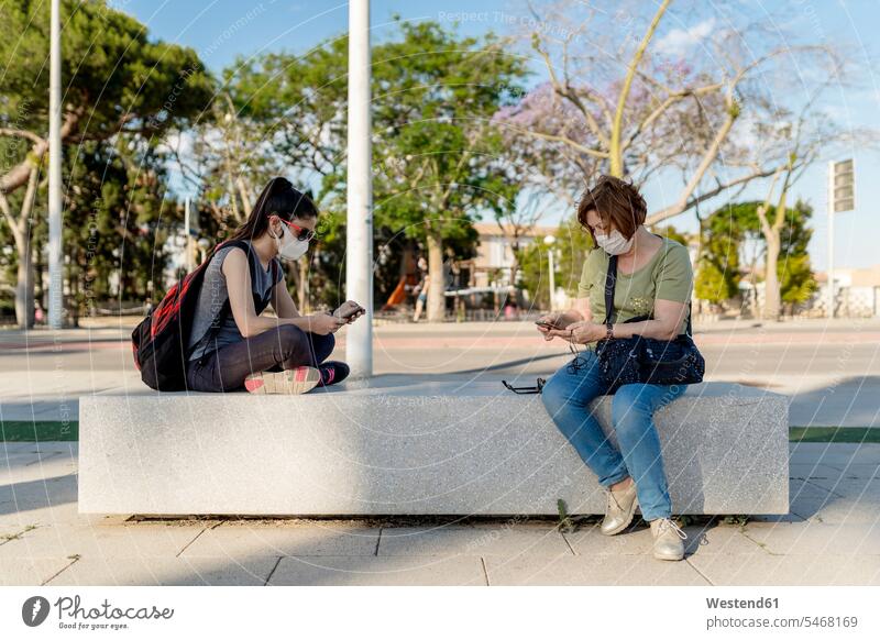 Frauen, die Masken tragen und Smartphones benutzen, während sie auf einem Sitz im Park sitzen Farbaufnahme Farbe Farbfoto Farbphoto Spanien