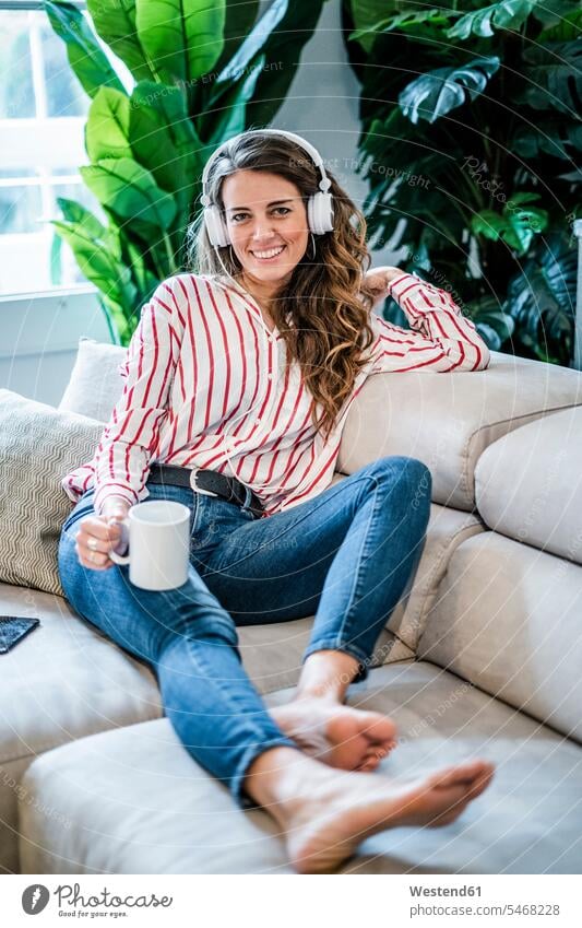Porträt der lächelnden Frau mit Tasse Kaffee und Kopfhörer sitzen auf Couch Portrait Porträts Portraits weiblich Frauen sitzend sitzt Kaffeetasse Kaffeetassen