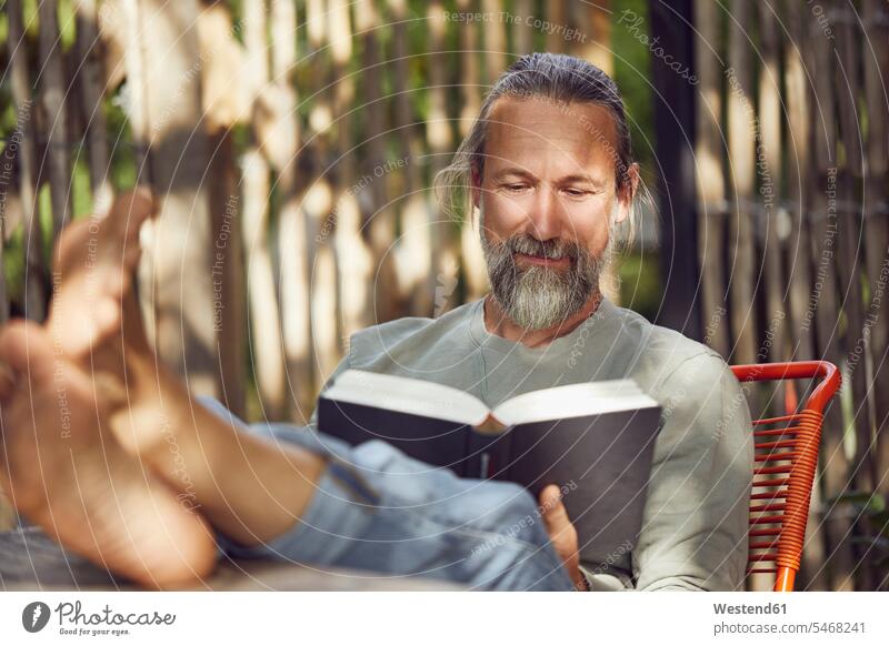 Bärtiger, reifer Mann liest Buch, während er sich auf einem Stuhl im Hof entspannt Farbaufnahme Farbe Farbfoto Farbphoto Deutschland Freizeitbeschäftigung Muße