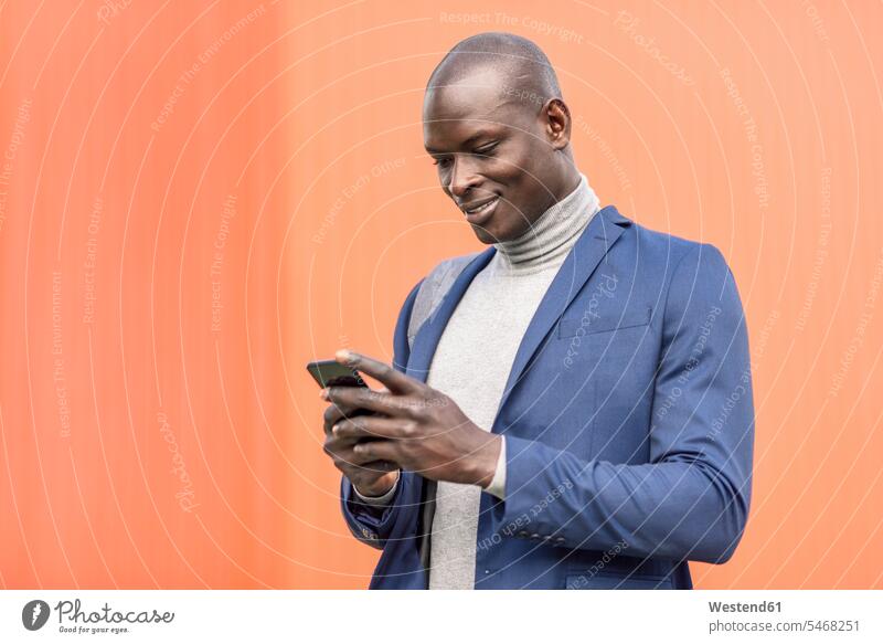 Lächelnder Geschäftsmann steht vor einer orangefarbenen Wand und schaut auf sein Smartphone iPhone Smartphones Wände Waende ansehen Portrait Porträts Portraits
