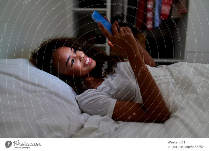Glückliche junge Frau mit Smartphone, während auf dem Bett im Schlafzimmer zu Hause liegen Farbaufnahme Farbe Farbfoto Farbphoto Innenaufnahme Innenaufnahmen