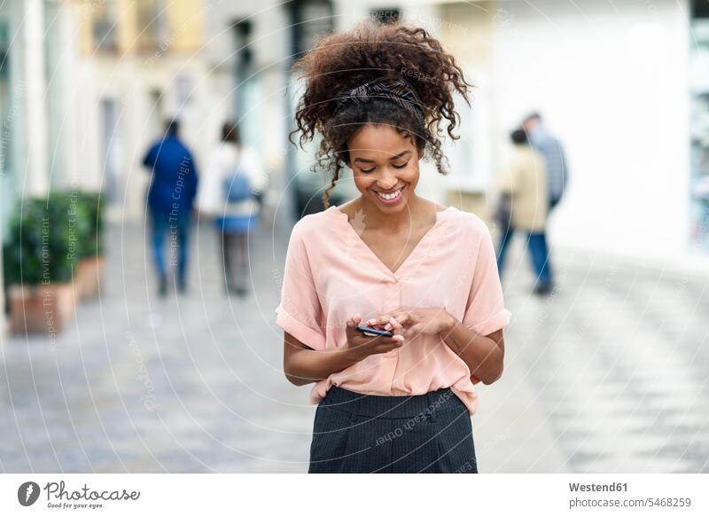 Lächelnde junge Frau mit Handy in der Stadt weiblich Frauen Mobiltelefon Handies Handys Mobiltelefone lächeln staedtisch städtisch Erwachsener erwachsen Mensch
