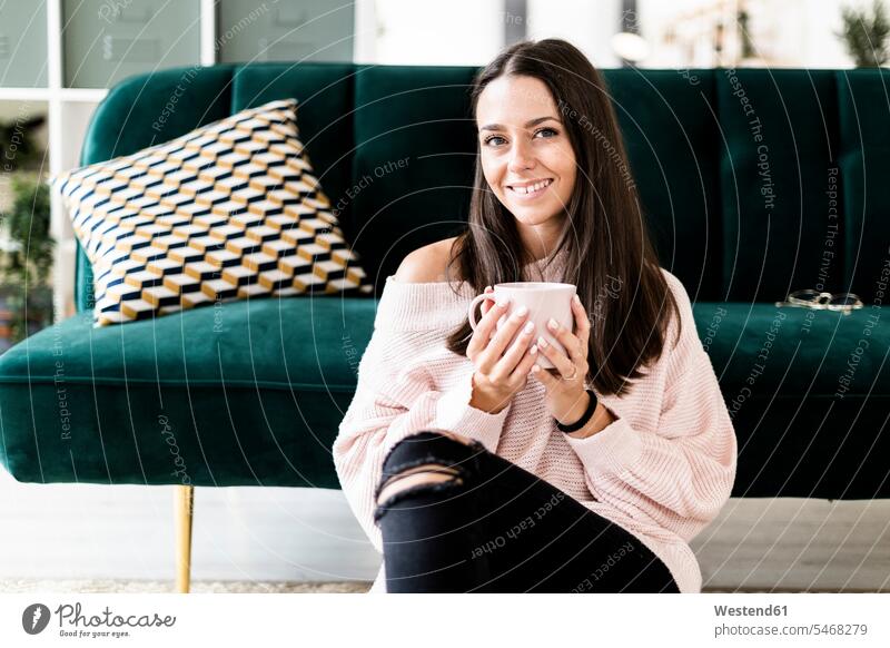 Lächelnde Frau sitzt mit Kaffeetasse vor dem Sofa zu Hause Farbaufnahme Farbe Farbfoto Farbphoto Innenaufnahme Innenaufnahmen innen drinnen Innenausstattung