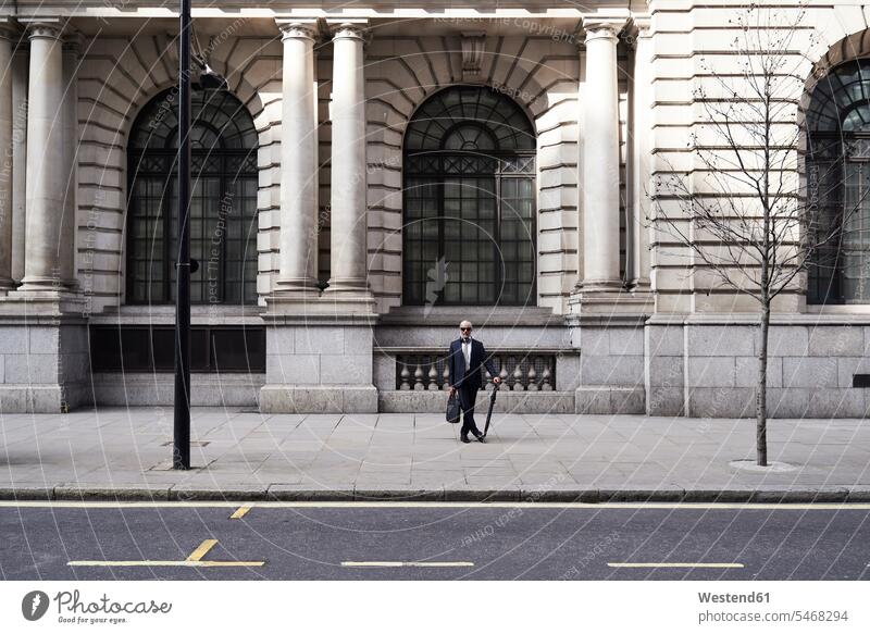 UK, London, stilvoller Senior-Geschäftsmann mit Aktentasche und Regenschirm auf dem Bürgersteig stehend Businessmann Businessmänner Geschäftsmänner Aktentaschen