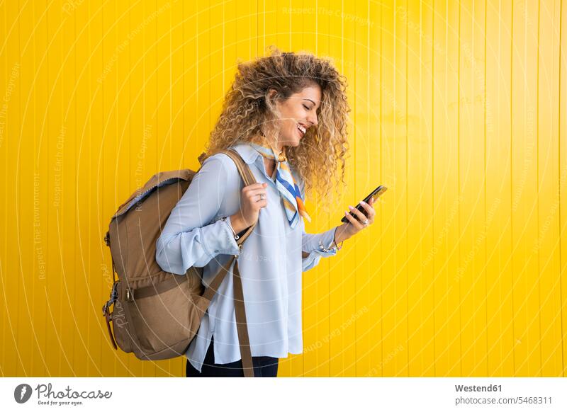 Lächelnde junge Frau mit Rucksack vor gelbem Hintergrund schaut auf Handy Leute Menschen People Person Personen Europäisch Kaukasier kaukasisch 1 Ein ein Mensch