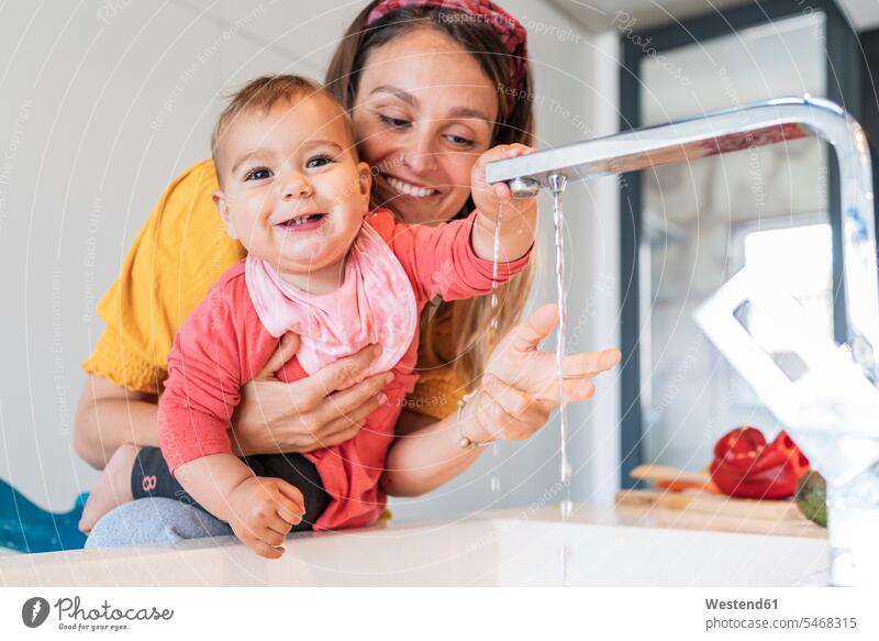 Lächelnde Mutter und süßes Mädchen spielen mit Wasser, das aus dem Wasserhahn in der Küchenspüle fällt Farbaufnahme Farbe Farbfoto Farbphoto Innenaufnahme
