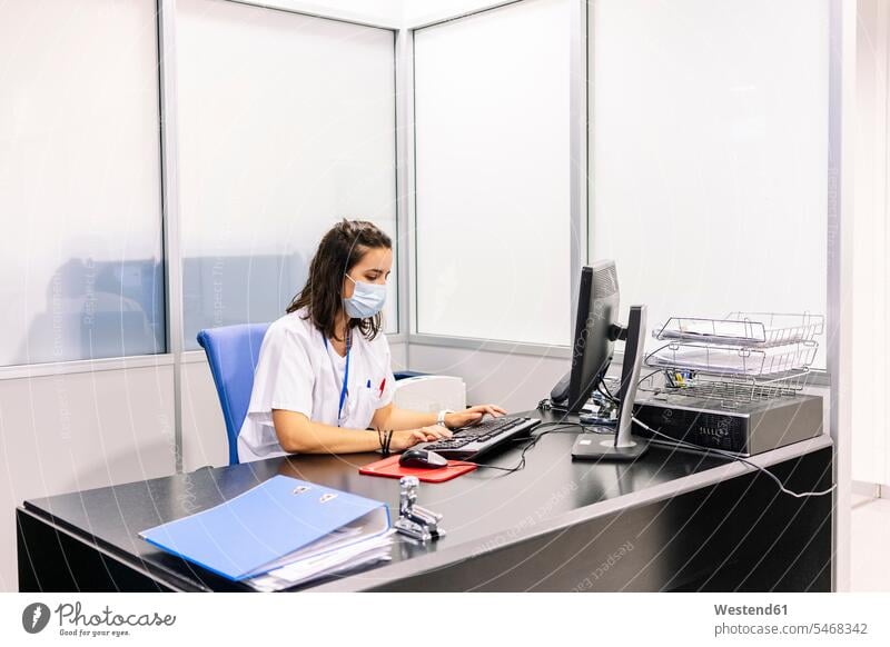 Ärztin mit chirurgischer Maske und Computer auf dem Schreibtisch im Büro im Krankenhaus Farbaufnahme Farbe Farbfoto Farbphoto Spanien Innenaufnahme