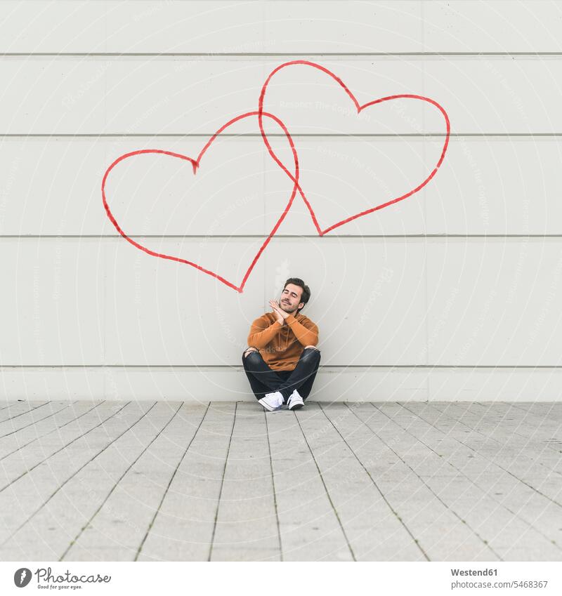Digitales Komposit eines jungen Mannes, der an einer Wand mit Herzen sitzt Leute Menschen People Person Personen Alleinstehende Alleinstehender Singles