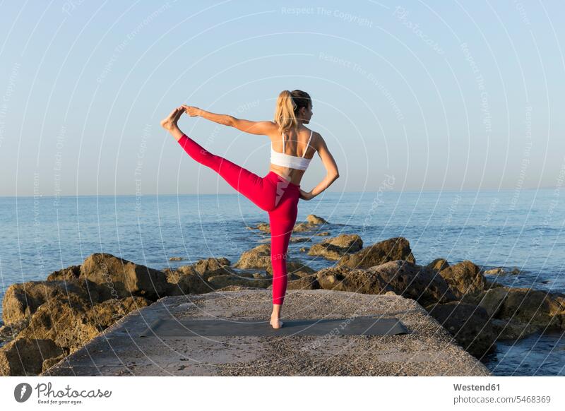 Junge Frau übt Yoga am Strand, macht stehende Balance, Utthita Hasta Padangustasana ausgeglichen Ausgeglichenheit Gleichgewicht ausüben trainieren Übung früh