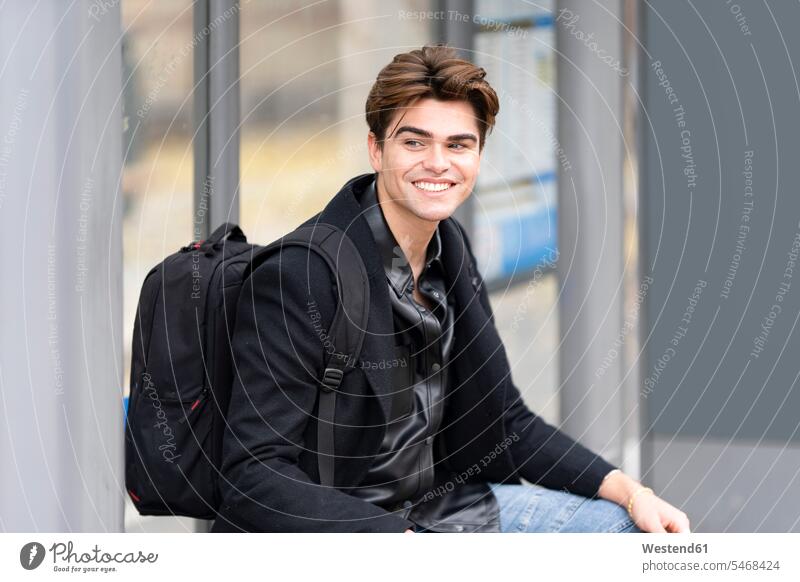 Lächelnder gut aussehender junger Mann, der wegschaut, während er mit Rucksack an der Bushaltestelle sitzt Farbaufnahme Farbe Farbfoto Farbphoto Spanien