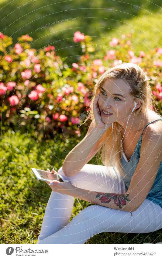 Lächelnde junge Frau sitzt im Park mit Handy und Ohrstöpsel Mobiltelefon Handies Handys Mobiltelefone Ohrhörer weiblich Frauen sitzen sitzend lächeln