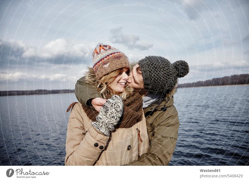 Junger Mann küsst seine Freundin vor einem See Handschuhe Kuss Küsse Jahreszeiten winterlich Winterzeit Glück glücklich sein glücklichsein gefühlvoll Emotionen