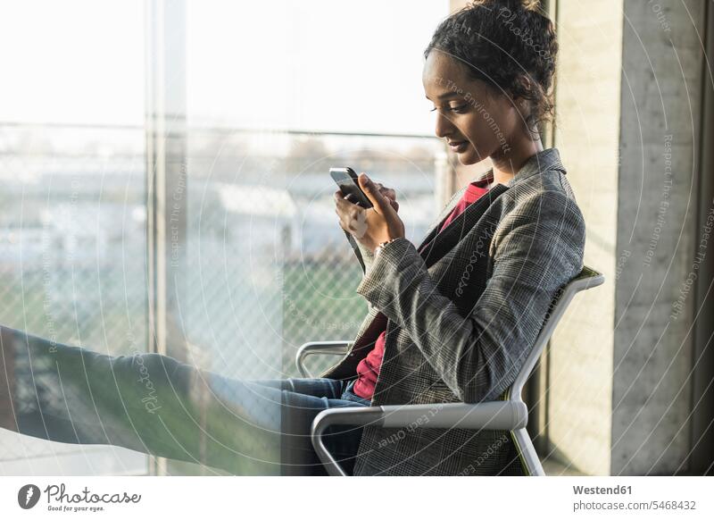 Junge Geschäftsfrau benutzt Handy am Fenster im Büro Job Berufe Berufstätigkeit Beschäftigung Jobs Leute Menschen People Person Personen gelockt gelockte Haare