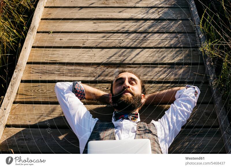 Gut gekleideter Mann mit Laptop liegt auf einem Holzsteg in der Landschaft Leute Menschen People Person Personen Europäisch Kaukasier kaukasisch 1 Ein