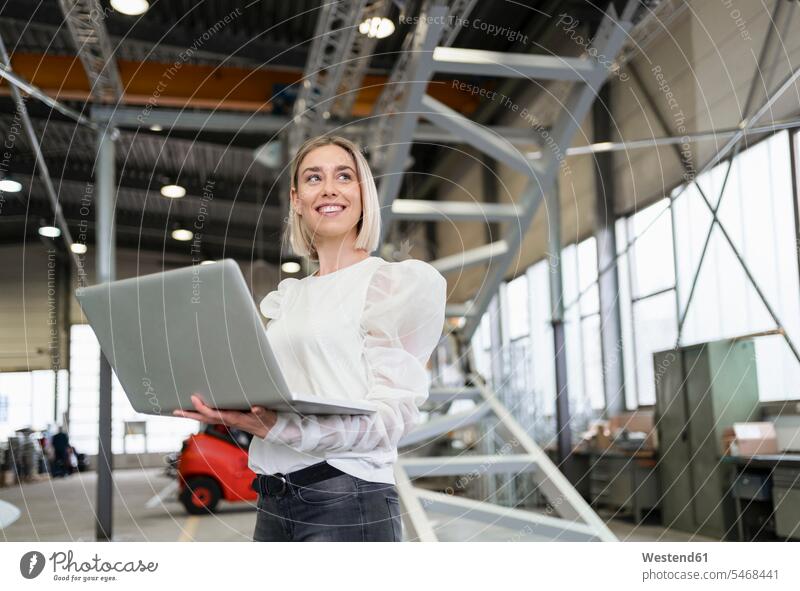 Lächelnde junge Frau mit Laptop in einer Fabrik Leute Menschen People Person Personen Europäisch Kaukasier kaukasisch 1 Ein ein Mensch nur eine Person single