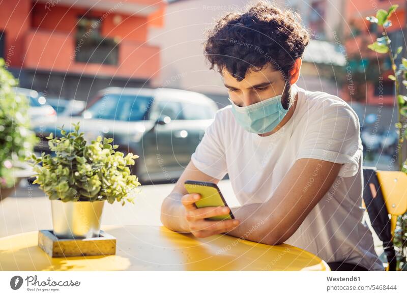 Junger Mann trägt Gesichtsmaske und benutzt ein Smartphone, während er im Straßencafé sitzt Farbaufnahme Farbe Farbfoto Farbphoto Freizeitbeschäftigung Muße
