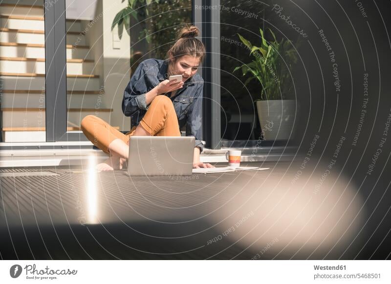 Junge Frau sitzt auf der Terrasse zu Hause mit Handy und Laptop Deutschland Freiberufler freiberuflich freie Berufe Zuhause arbeiten von zuhause arbeiten