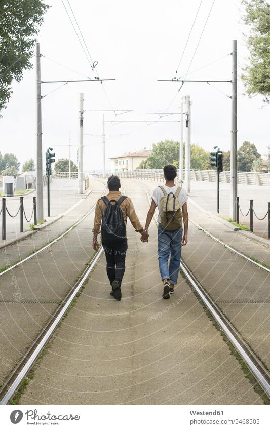 Rückenansicht von jungen Homosexuell Paar mit Rucksäcken zu Fuß Hand in Hand zwischen Spuren auf der Straße gehen gehend geht Pärchen Paare Partnerschaft schwul
