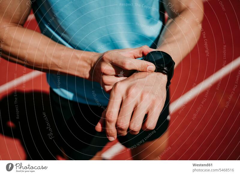 Männlicher Athlet auf Tartanbahn bei der Kontrolle von Smartwatch Uhren Armbanduhren Farben Farbtoene Farbton Farbtöne rote roter rotes stehend steht Muße