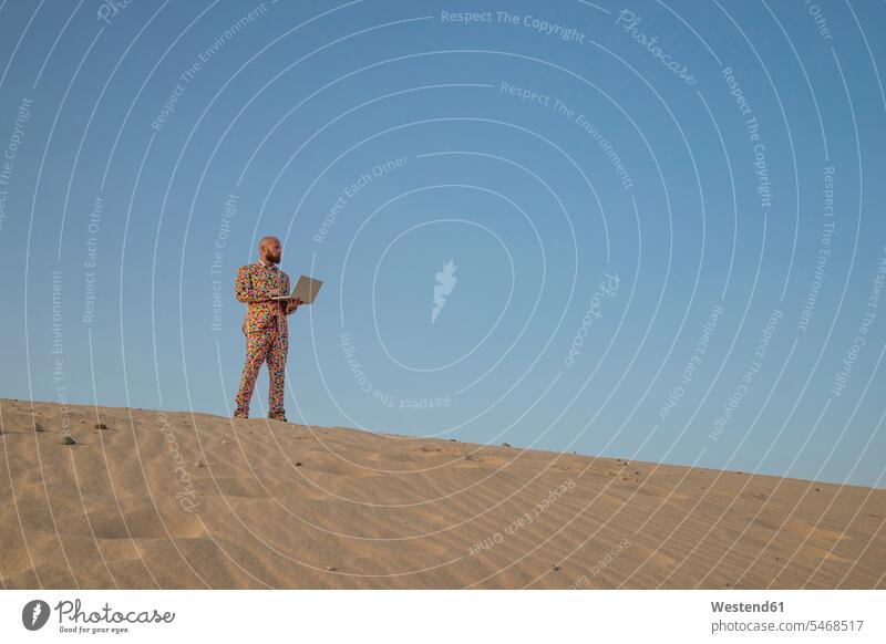 Mann mit Laptop im Anzug mit bunten Tupfen steht auf einer Sanddüne und schaut in die Ferne Sanddünen gepunktet getupft Punktmuster farbenfroh Weite Männer